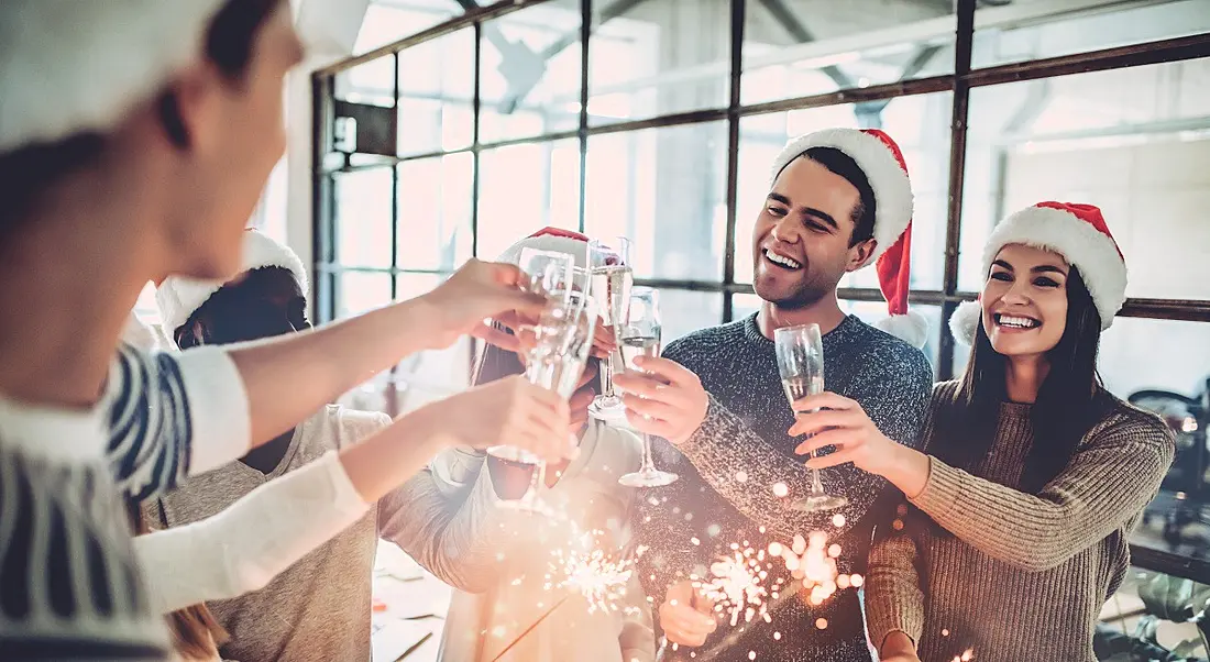 A diverse group of employees wearing Santa hats and clinking champagne glasses at the office Christmas party.