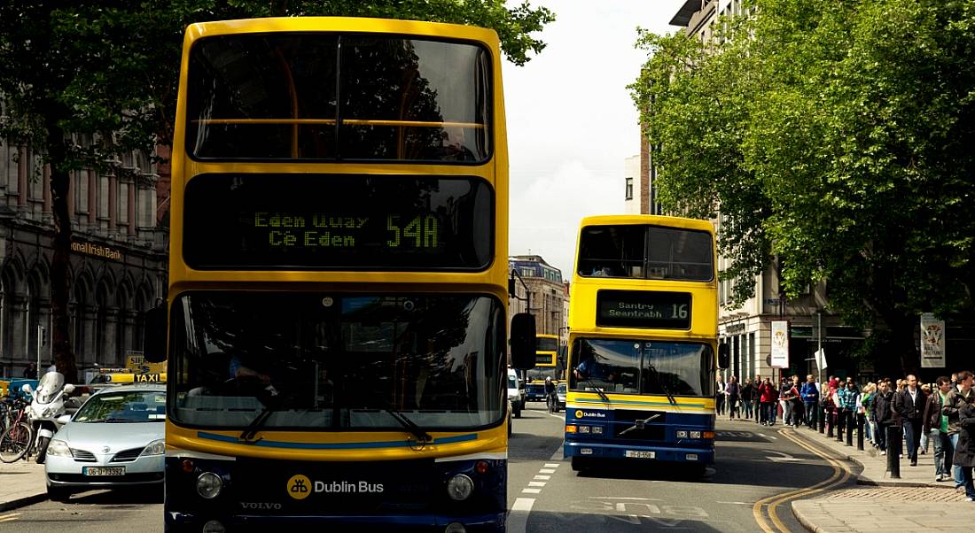 Two Dublin buses driving down a main street in Dublin city on a bright, clear, cloudless day.