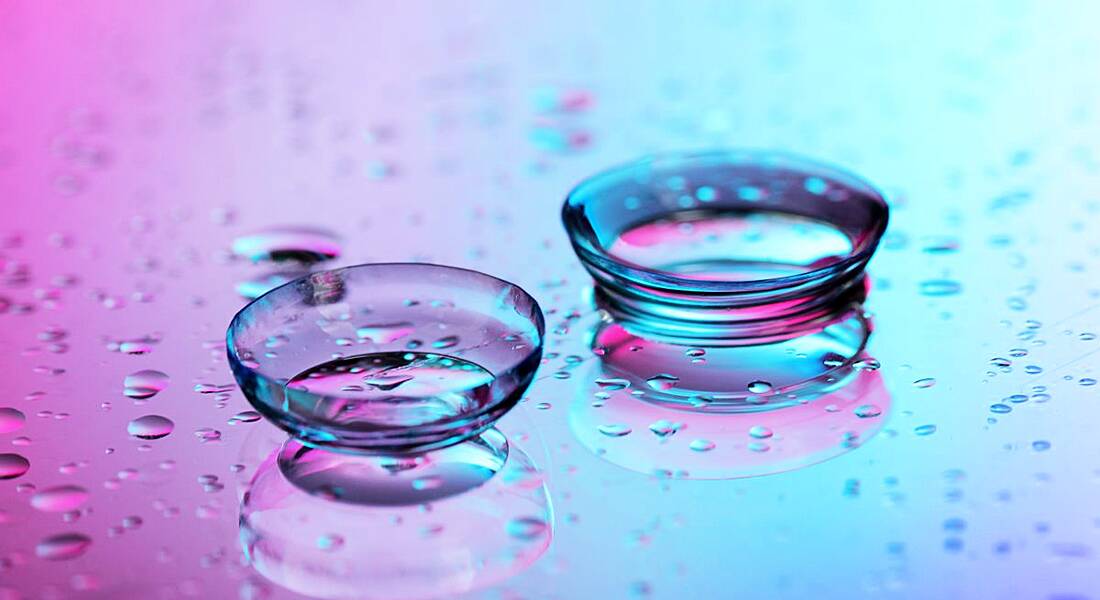 Contact lenses surrounded by water droplets on pink-blue background.