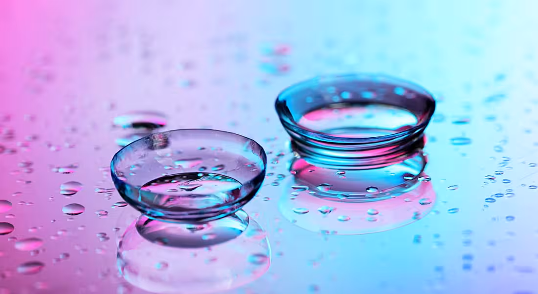 Contact lenses surrounded by water droplets on pink-blue background.