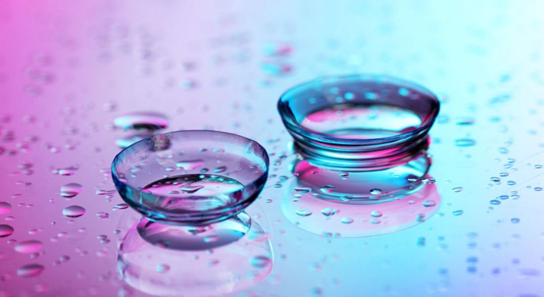 Contact lenses surrounded by water droplets on pink-blue background.