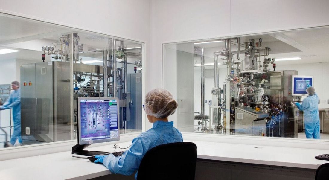 A woman with a science job in a blue lab coat sitting at a laptop in a lab office. Other scientists work in the background.