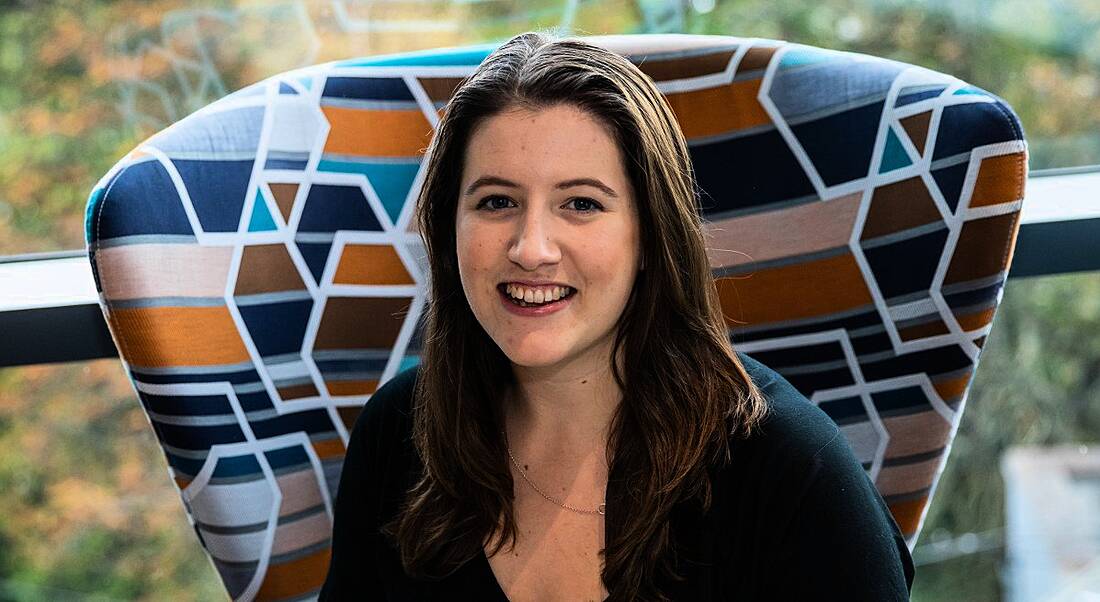 A brunette woman with long flowing hair sitting on a chair with a geometric pattern smiling against the camera against a window.