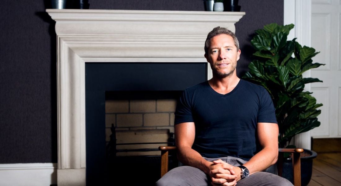 man with his arms held in his lap sitting and looking into the camera against the backdrop of a navy wall, sprightly potted ferm and white fireplace.