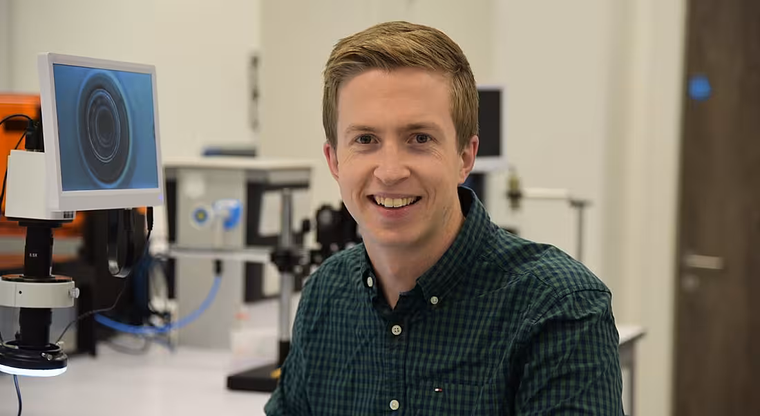 A smiling fair-haired young man in a check shirt with a medtech lab in the background.