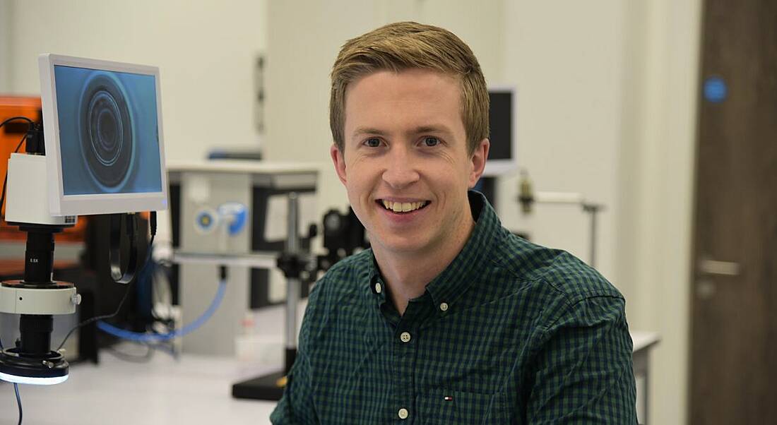 A smiling fair-haired young man in a check shirt with a medtech lab in the background.