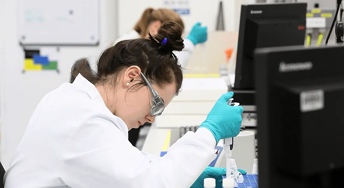 A young female scientist in blue visor glasses hunched over pharmaceutical equipment.