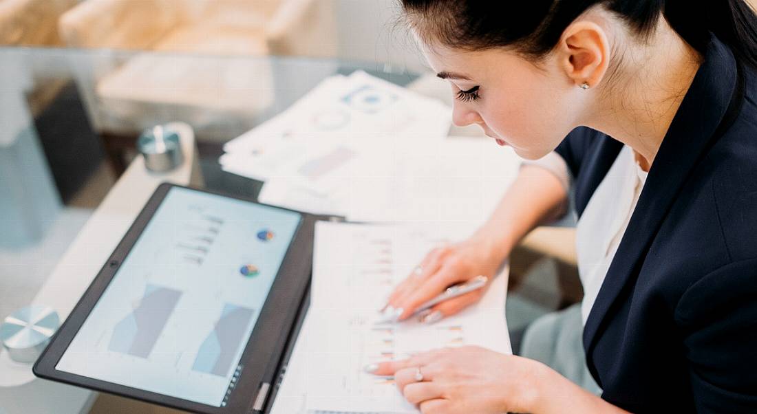 woman with dark hair looking at papers in front of a tablet with graphs and pie charts.