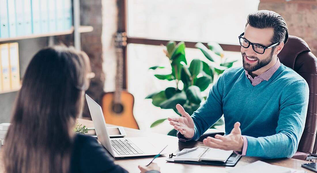 A smiling man is sitting at a desk. A woman sits opposite him. He is interviewing her for a DevOps job.