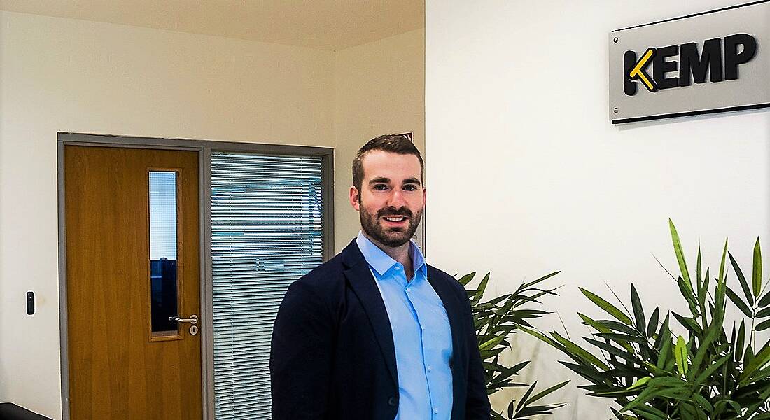 A young bearded man in a blue shirt and navy blazer giving a slight smile. A Kemp logo can be seen on the wall behind him.