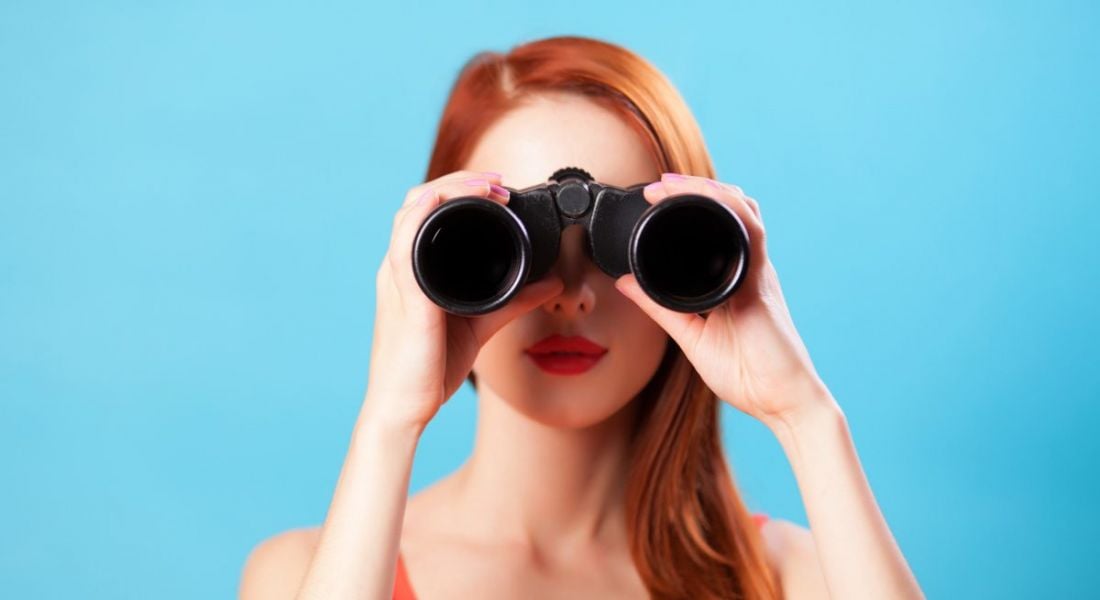 A young female jobseeker with red hair looking through a pair of binoculars against a blue background.