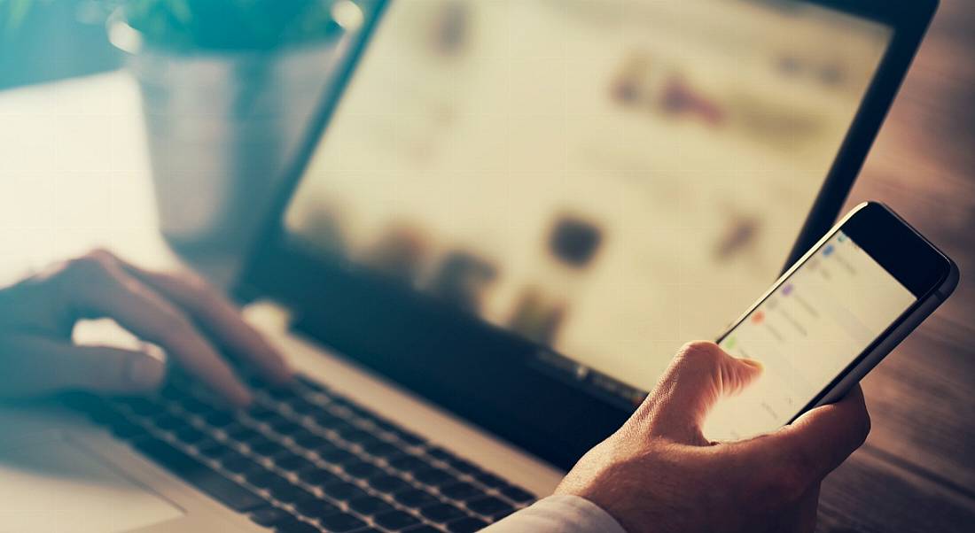 View of a man’s hand and upper arm holding a phone with an open laptop in the background.