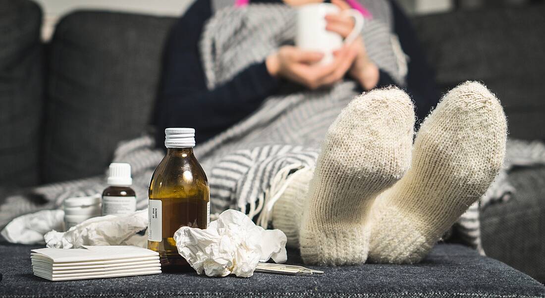 View of sock-clad feet of someone who is sick surrounded by crumpled tissues and cough syrup bottles.