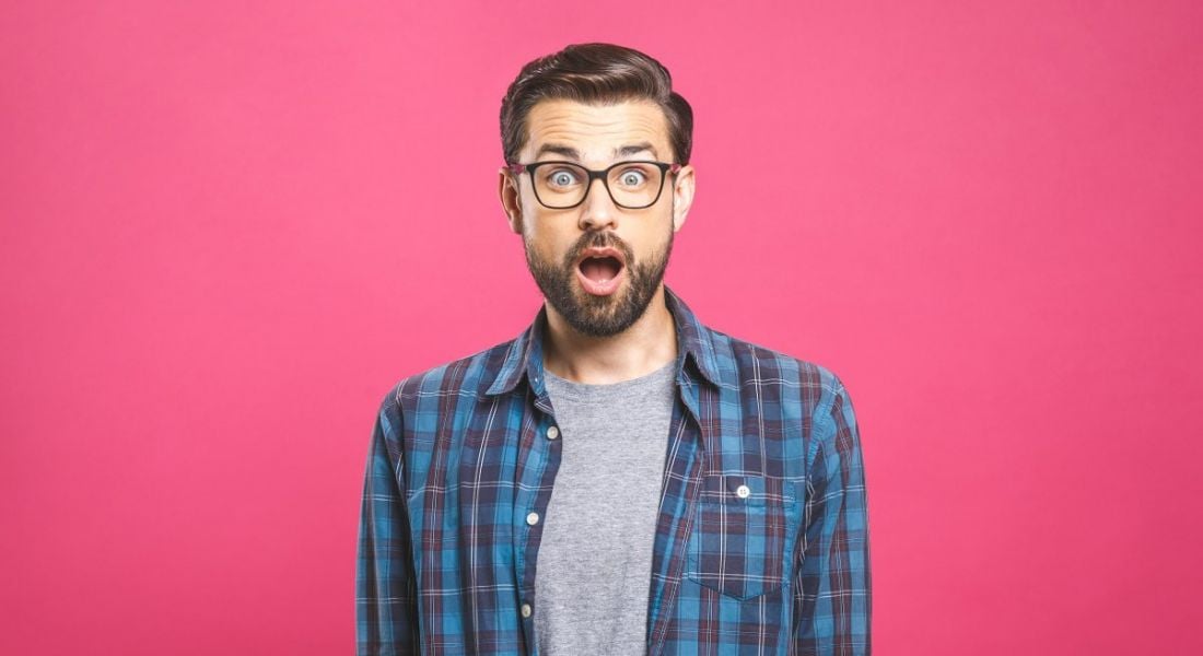 man with glasses and plaid shirt looking shocked, his jaw slack, against a fuschia pink background.