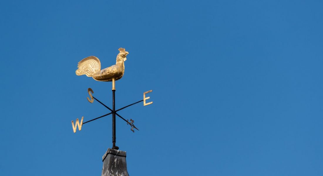 Image of a slightly tarnished gold weather vane with a rooster in the centre facing east against a cloudless blue sky.