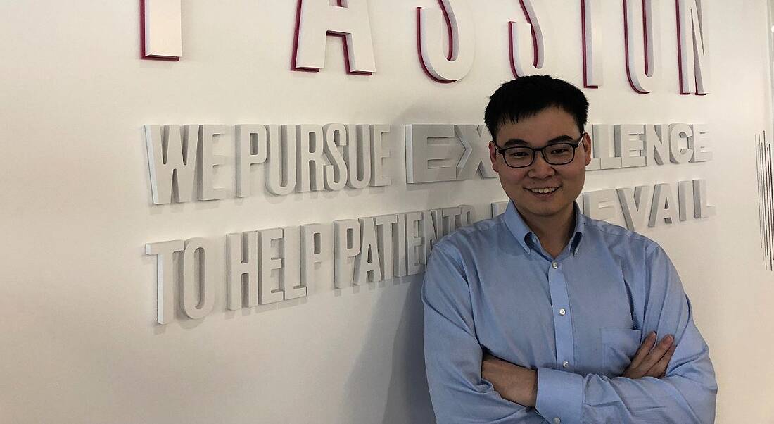 A young man with thick-rimmed glasses and a powder blue shirt smiling and leaning against a beige wall.