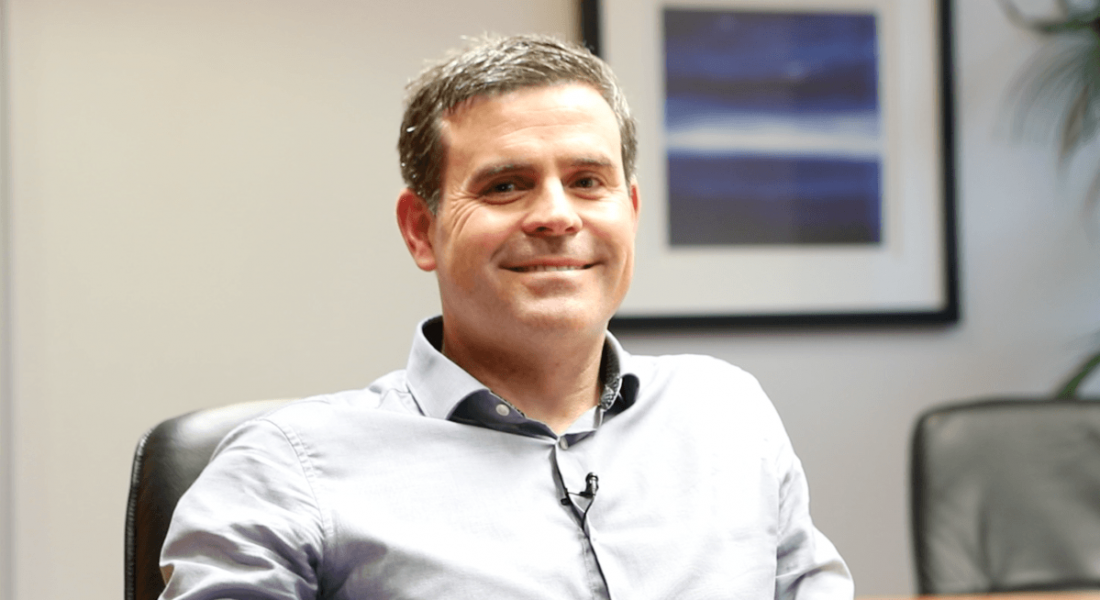 A smiling professional man in an open-neck light grey shirt.