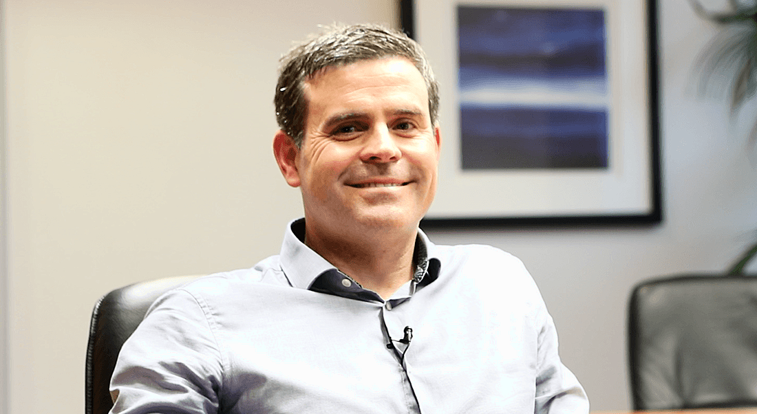 A smiling professional man in an open-neck light grey shirt.