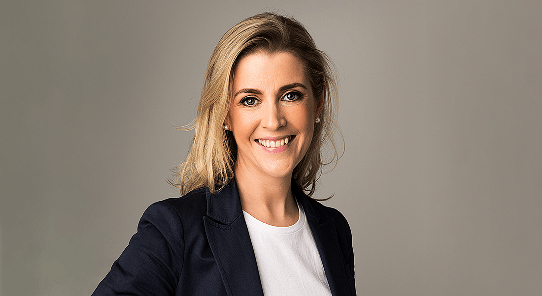 A blonde woman smiling at the camera against a beige background. She’s wearing a navy blazer and white t-shirt.