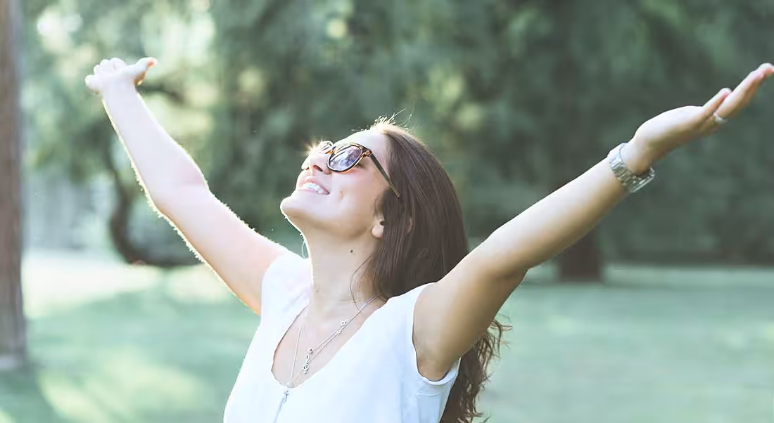 A smiling woman standing outside with arms outstretched to the sky. She has a very positive mindset.