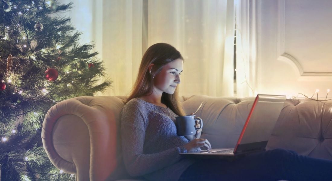 A woman lying on a couch working on a laptop and holding a mug. Behind her is a Christmas tree. She’s hoping to find a job.