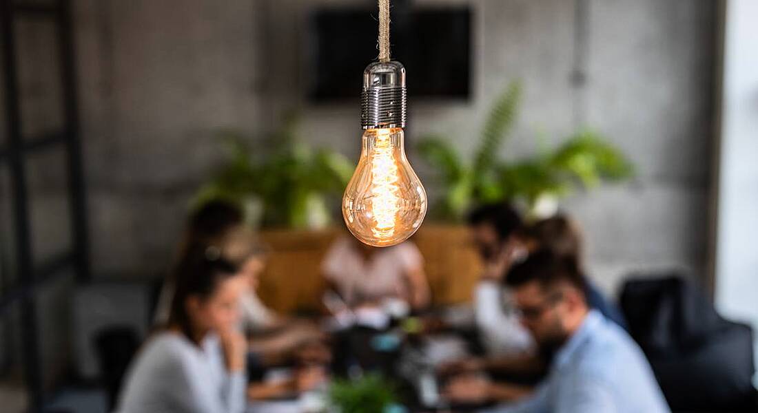 exposed lightbulb in focus in centre foreground with group of young business people working in the background.