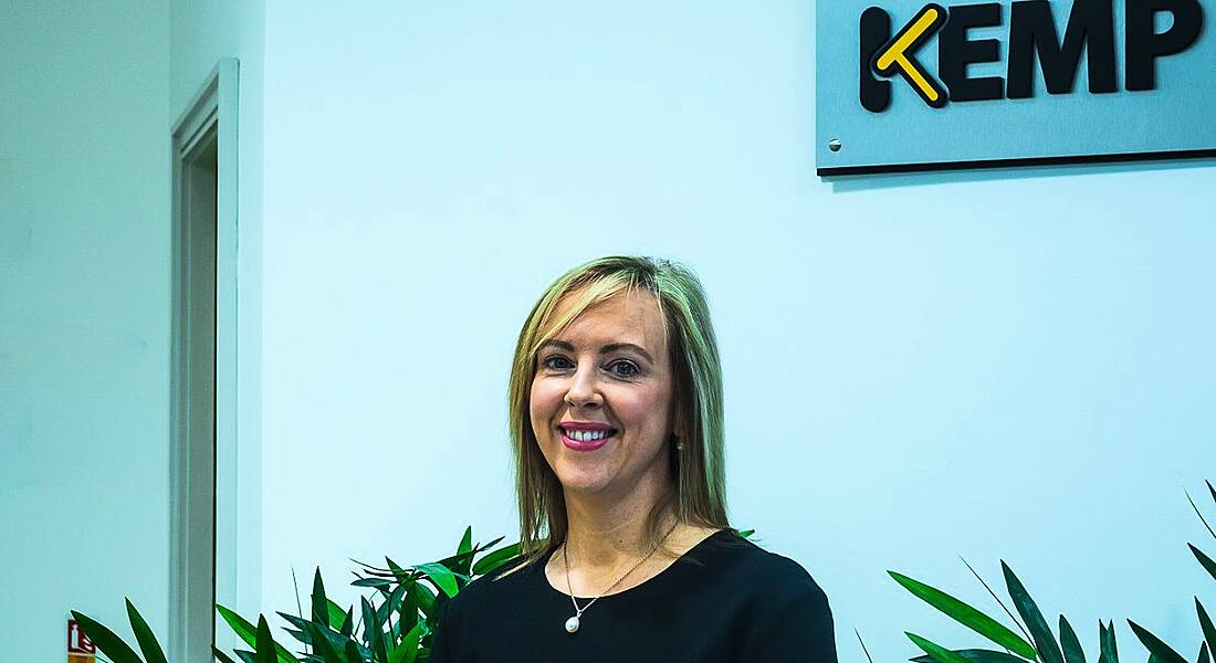 A blonde woman smiling in an office with the Kemp Technologies logo in the background on the wall.