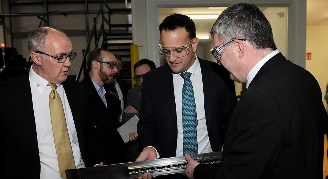 Three suited men looking down at and inspecting a long, thin titanium aircraft part.