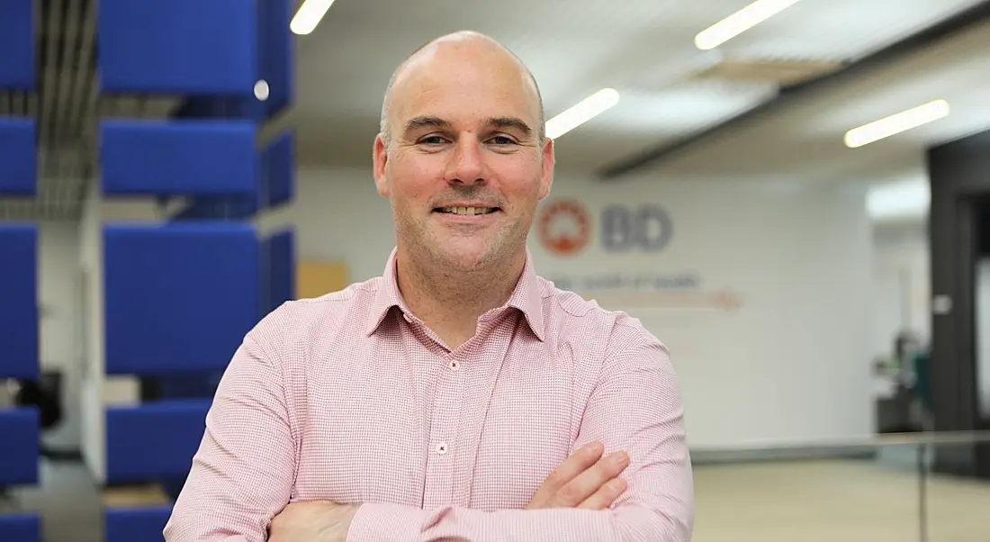 A smiling, middle-aged bald man in a light pink shirt standing in BD. The BD logo is on the wall behind him.