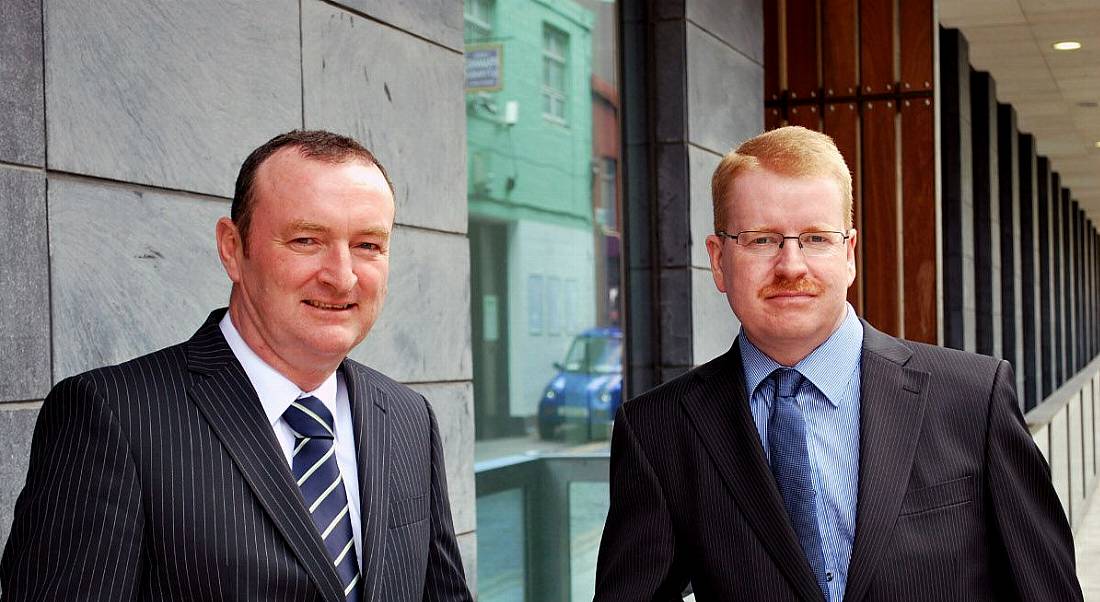 One dark-haired man and one red-haired man in suits stand outside a building.