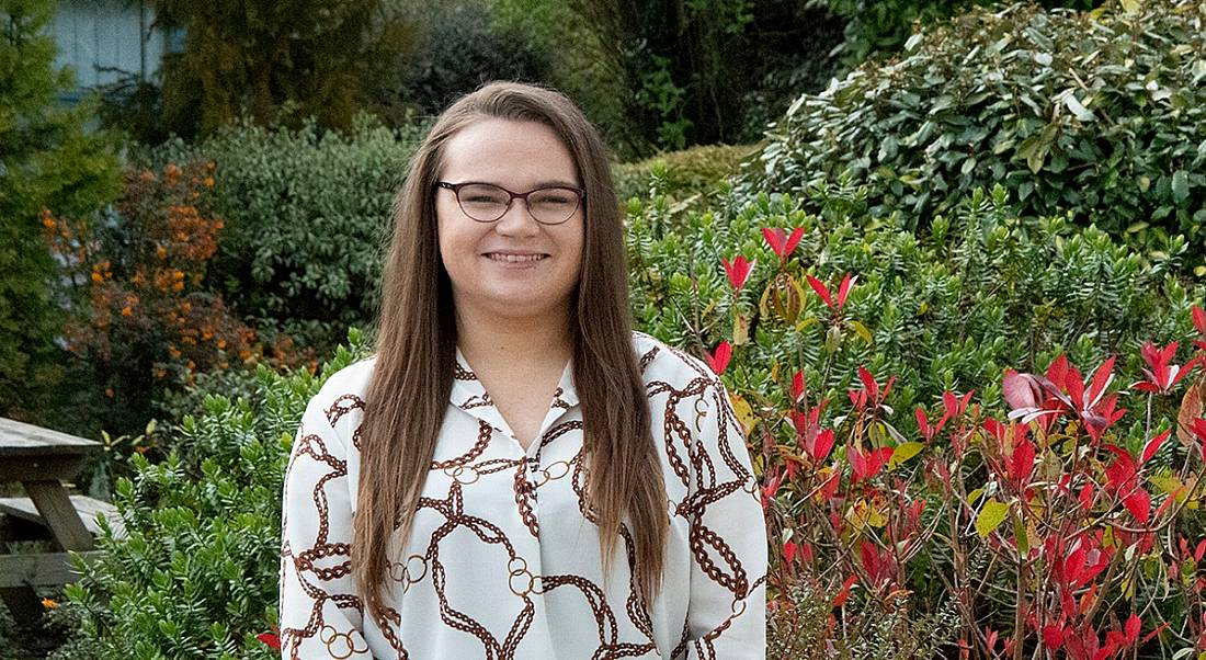 A young woman in a white patterned shirt with glasses and long brown hair standing in front of greenery outside.