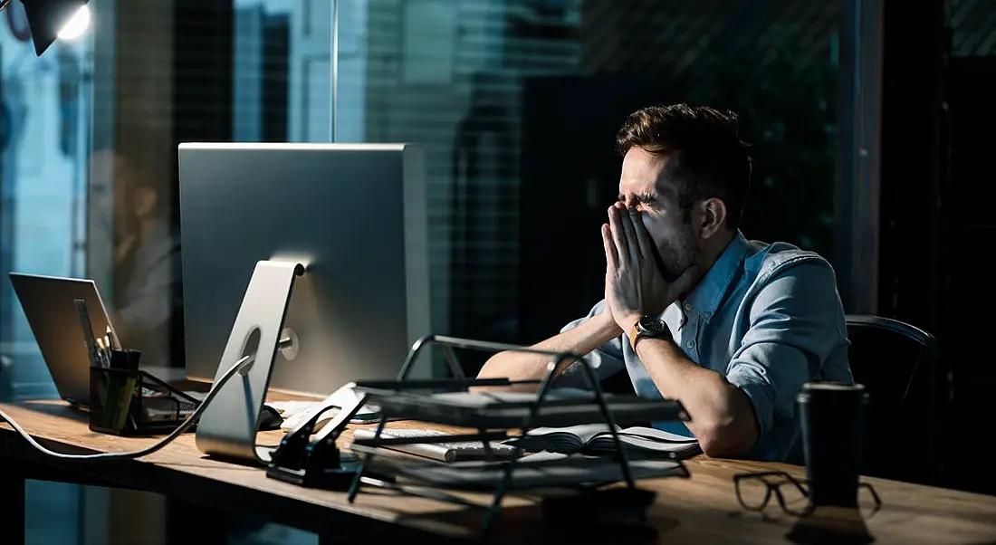 A man at a desk in a dark office covering his face in frustration. He is overworking and feeling burnt out.