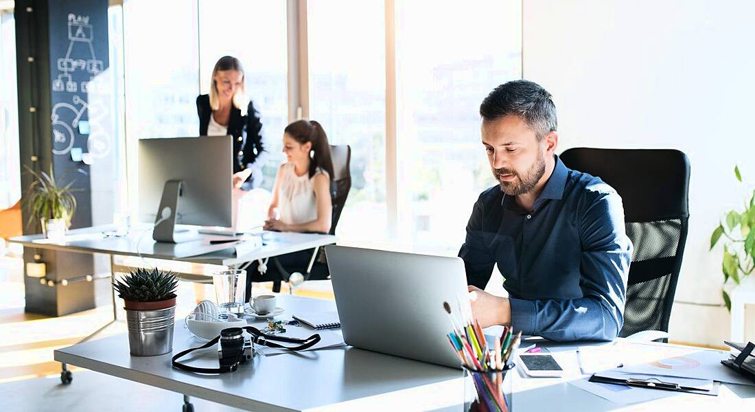 A man at a desk in a bright office with two female workers in the background. They are having a good employee experience.