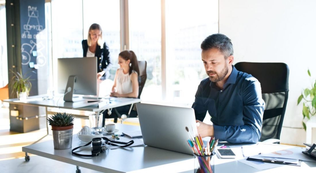 A man at a desk in a bright office with two female workers in the background. They are having a good employee experience.