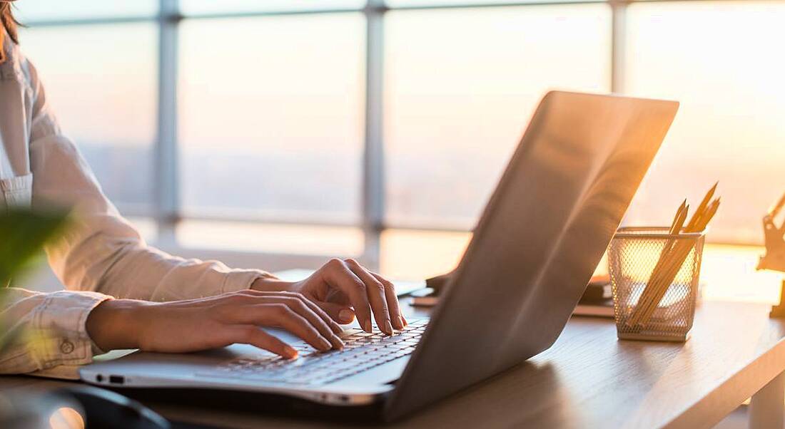 View of a desk lit by sunlight streaming in from a nearby window, with a woman's hands typing at a laptop.