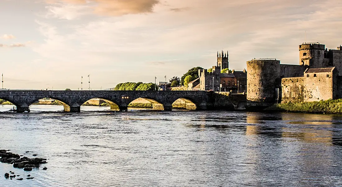 A picturesque view of Limerick from across a river with a bridge and a castle in the background.