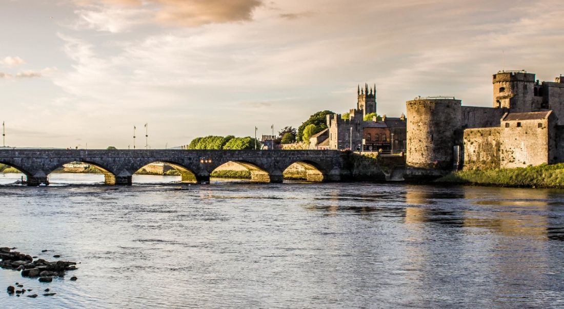 A picturesque view of Limerick from across a river with a bridge and a castle in the background.