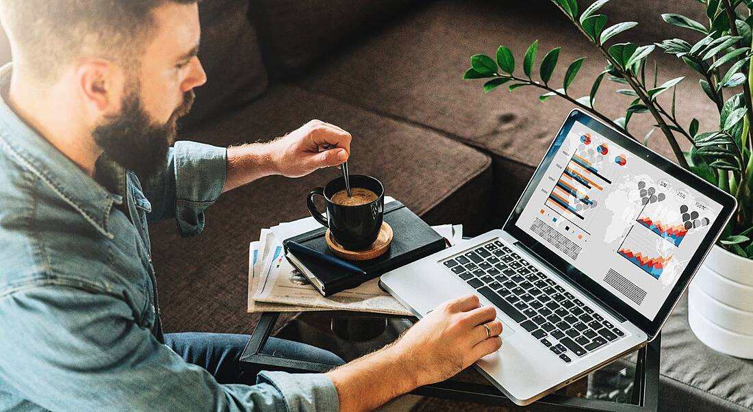 Young bearded man in blue shirt stirring a coffee with his left hand and using a laptop with the right.