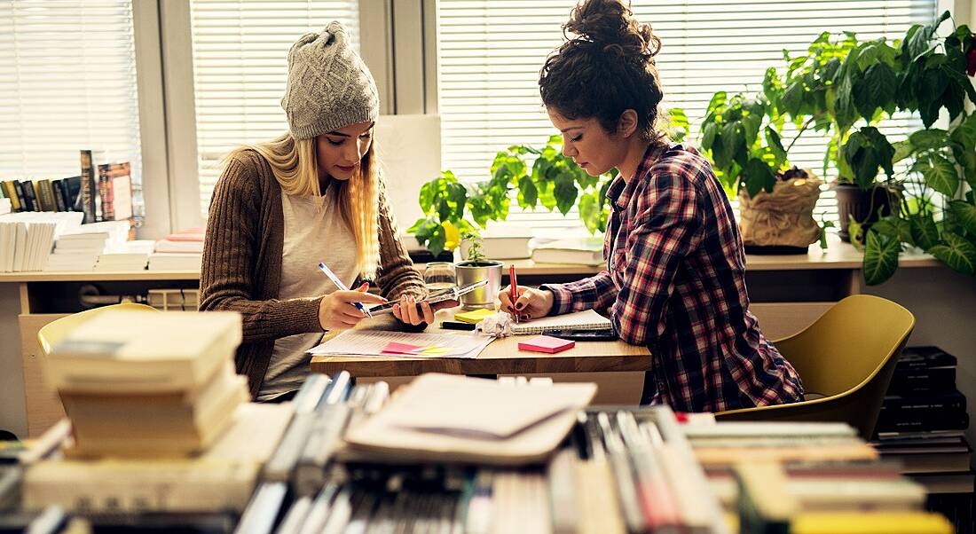 View of two women at a small desk holding highlighters and pens poring thoughtfully over their CVs surrounded by stacks of books.