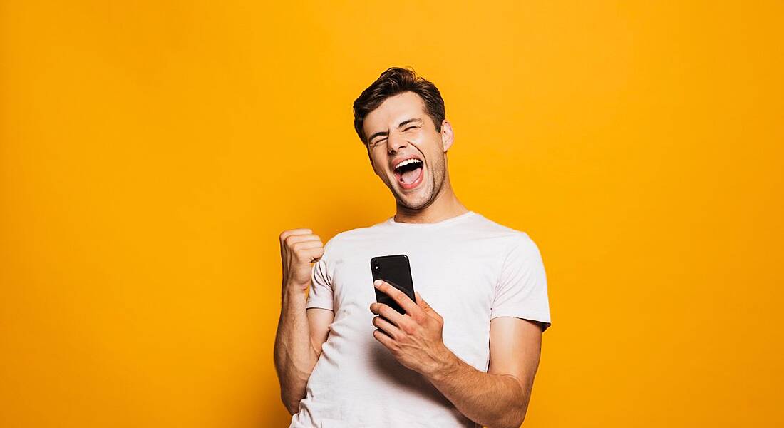 Young man in white t-shirt holding phone with joyful expression on his face against a mustard yellow background.