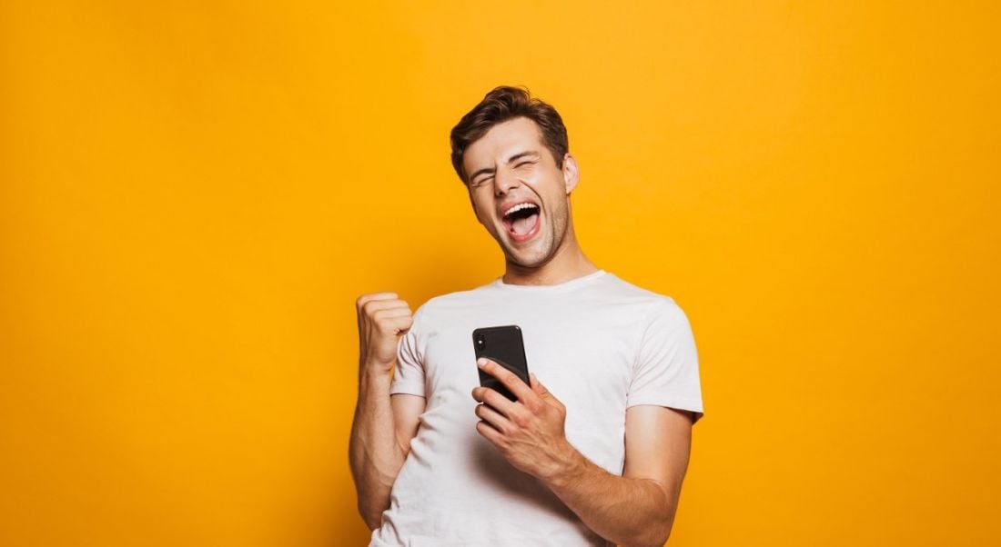 Young man in white t-shirt holding phone with joyful expression on his face against a mustard yellow background.