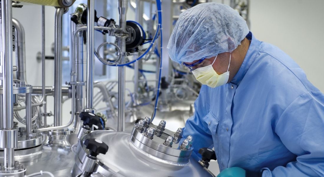 A man in a lab coat, mask and hair net in a biopharma manufacturing facility lab.