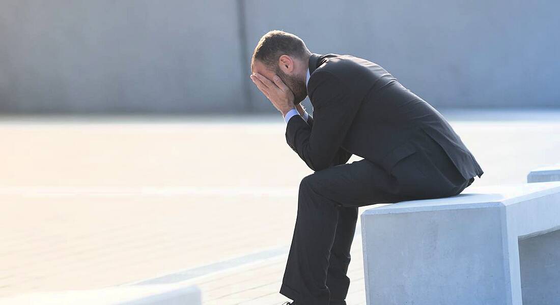 A man in a suit sitting on a bench outdoors with his head in his hands after a bad interview.