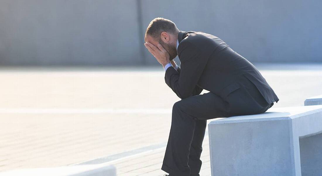 A man in a suit sitting on a bench outdoors with his head in his hands after a bad interview.