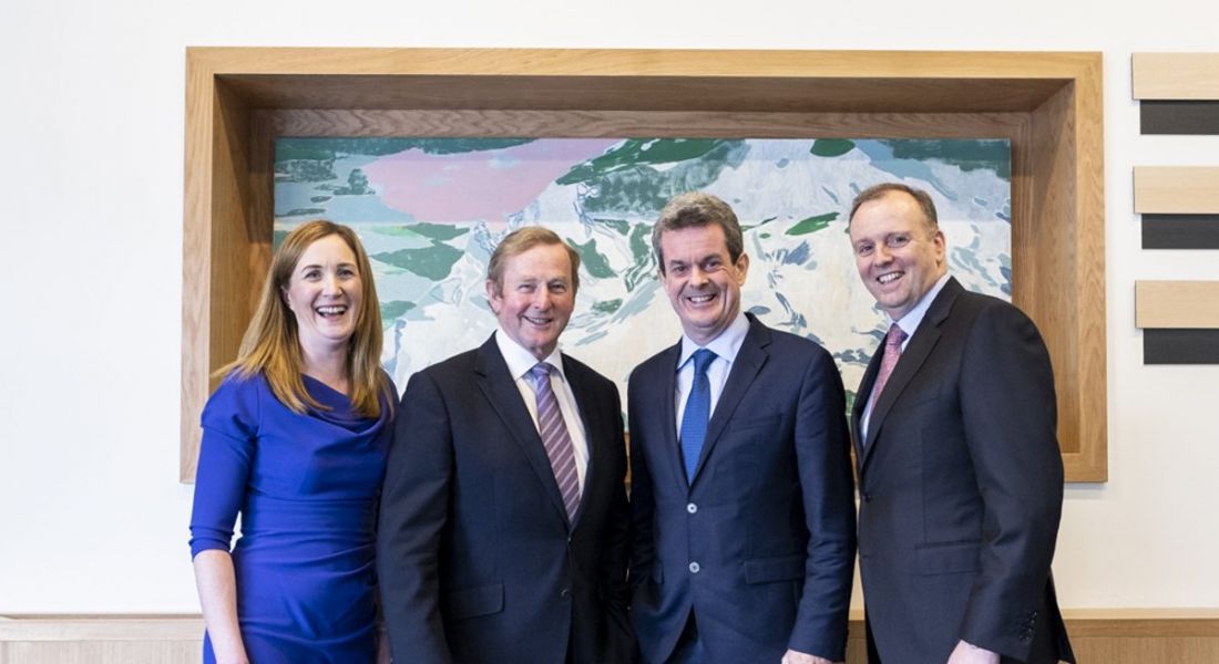 One woman and three men in business attire standing on a grey carpet in front of a painting and smiling at the camera.