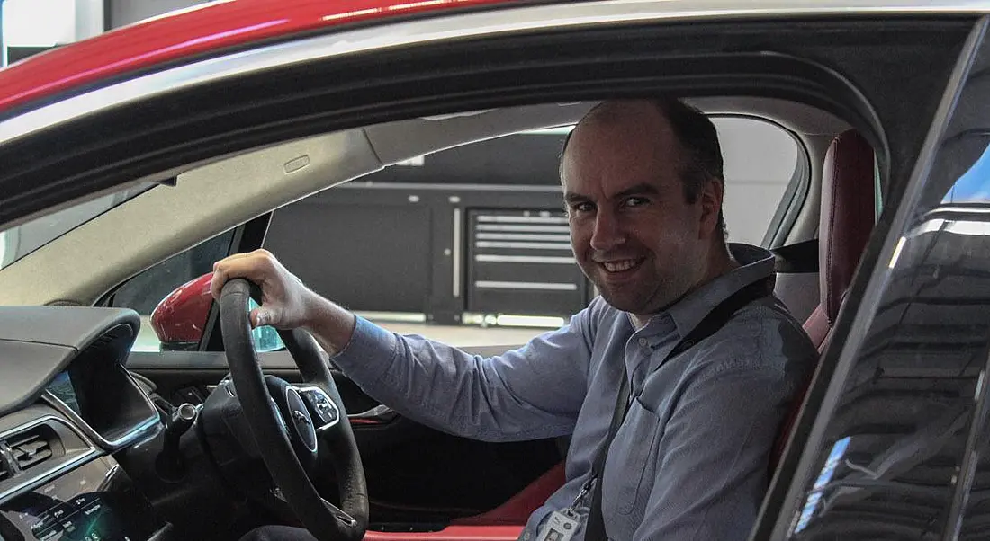 A man in a light blue shirt wearing a seatbelt in a car smiling at the camera.