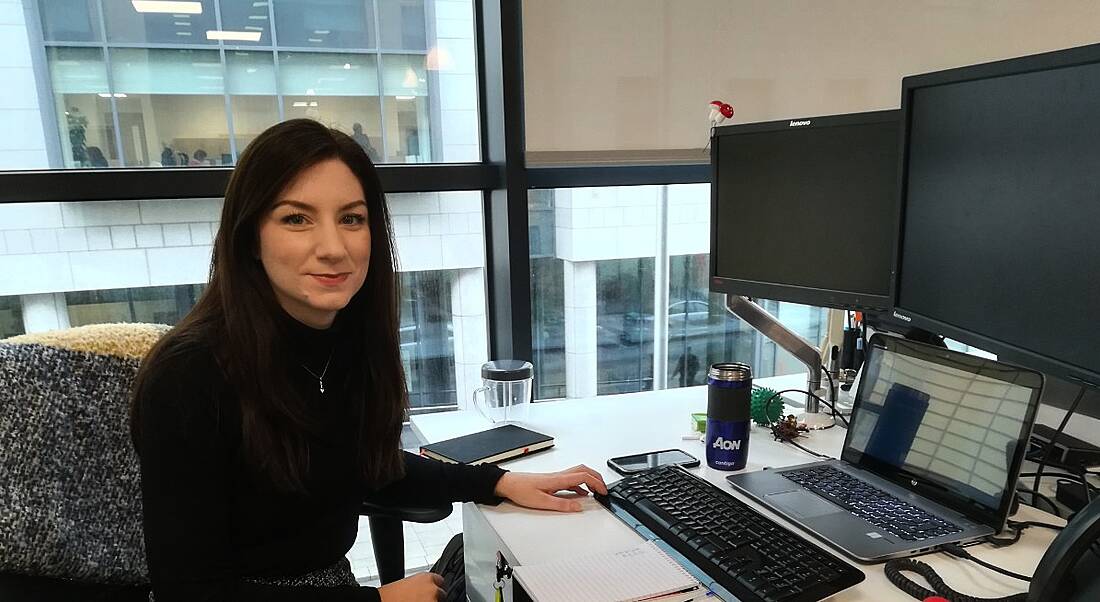 A young brunette woman in a black sweater sitting at an office desk with her chair turned towards the camera.