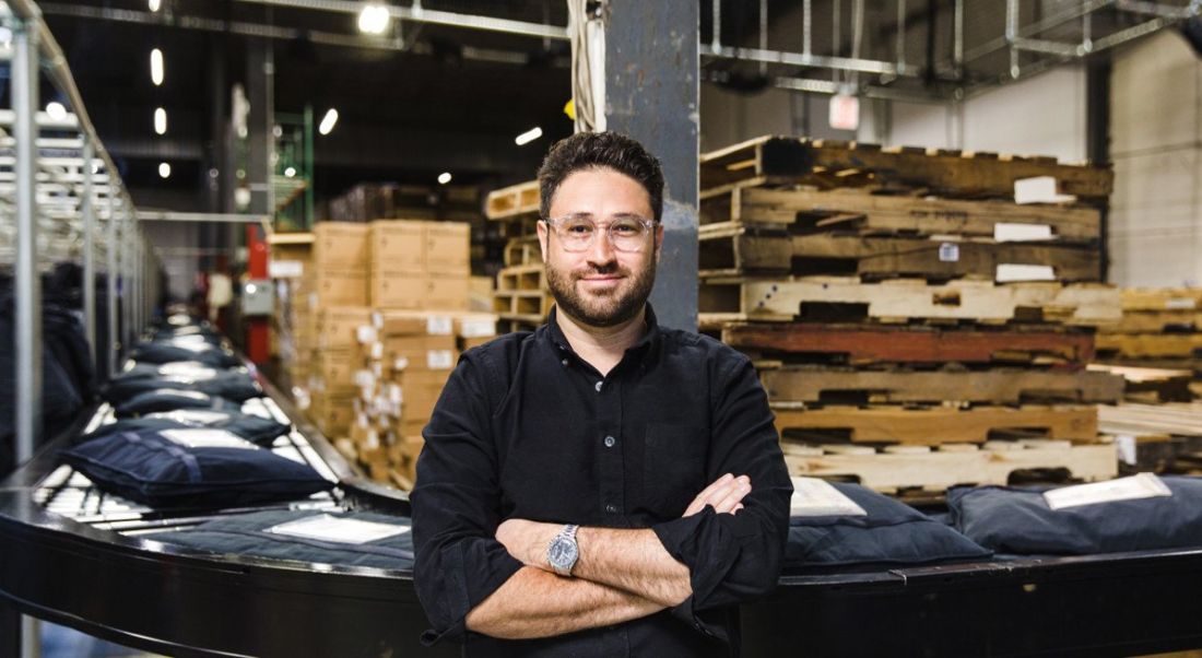 Man with dark hair, glasses and black shirt stands with arms folded in front of an assembly line with garments.