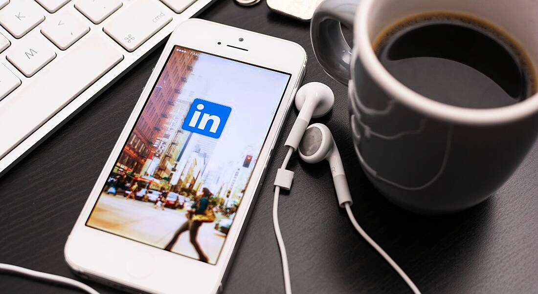 A close-up of a smartphone with a LinkedIn logo on the screen. The phone is on a table beside a coffee and earphones.