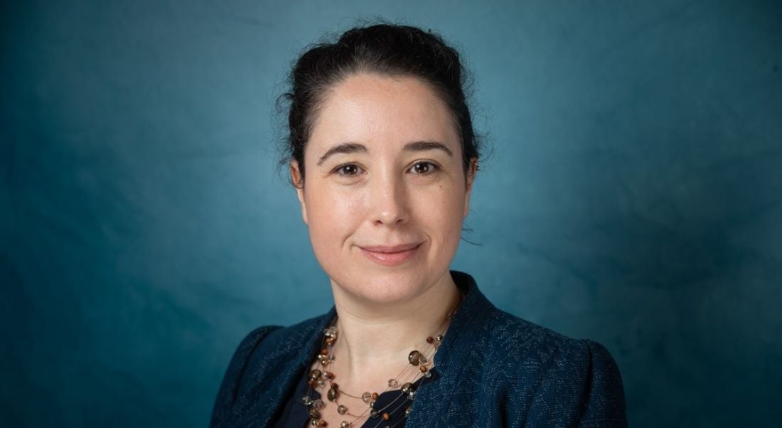 Dark haired woman wearing navy looking directly at the camera and smirking against aquamarine background.